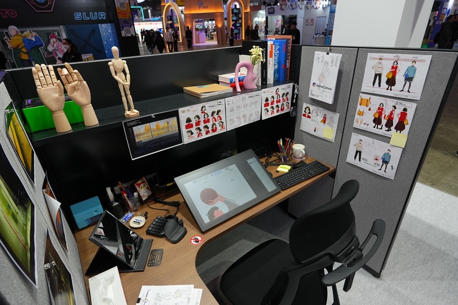 A display modeled after a creator’s desk, featuring a pen display tablet and keyboard, with a wooden artist’s mannequin, a hand model, and reference materials posted on the shelf.
