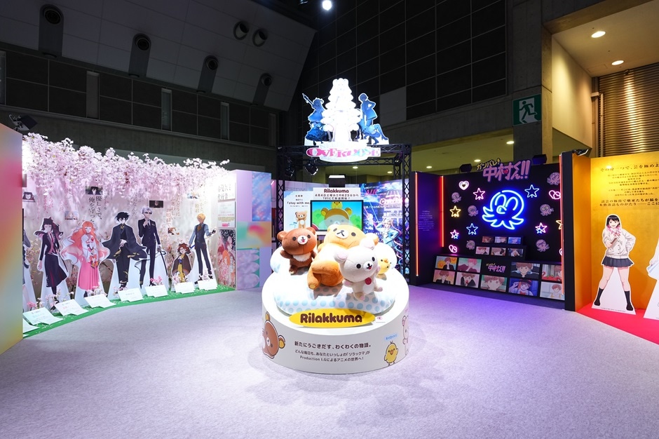 Panels from multiple anime works arranged in a curved layout, with a Rilakkuma plush displayed at the center.