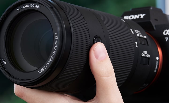 A close-up of a telephoto camera lens mounted on a mirrorless camera, with a hand pressing a control button on the lens barrel.