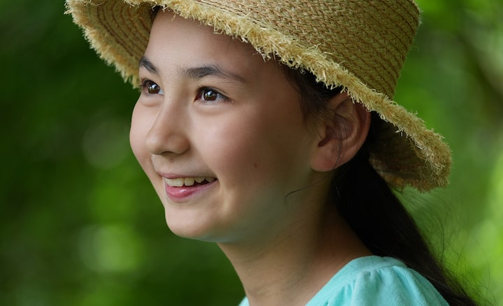 A smiling girl wearing a straw hat looks off into the distance, surrounded by soft green foliage in natural light.