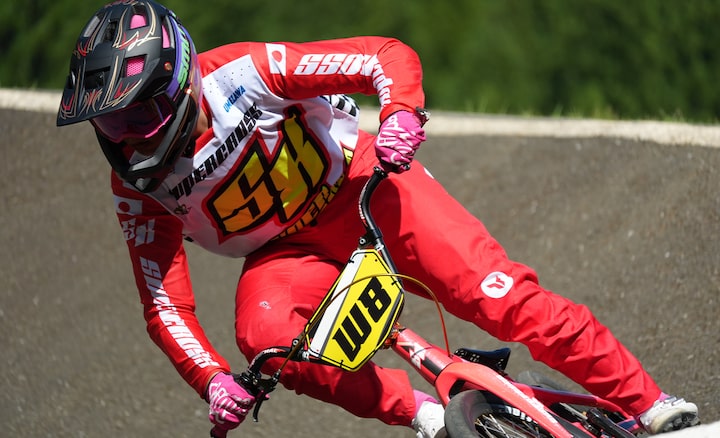 A BMX rider in bright red racing gear leans sharply into a turn on a dirt track during a high-speed race.