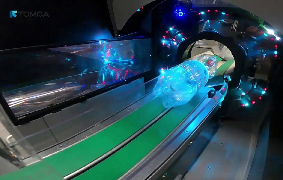 飲料容器自動回収機の画像。青く照らされたペットボトルが、計器のついた開口部からベルトコンベア沿いに機器内に入っていく。